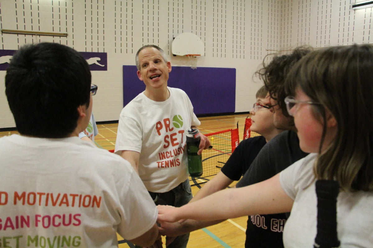 A group of people standing in a circle inside a gymnasium, wearing matching white t-shirts with red and green text that reads 'PRO SET INCLUDES TENNIS'.